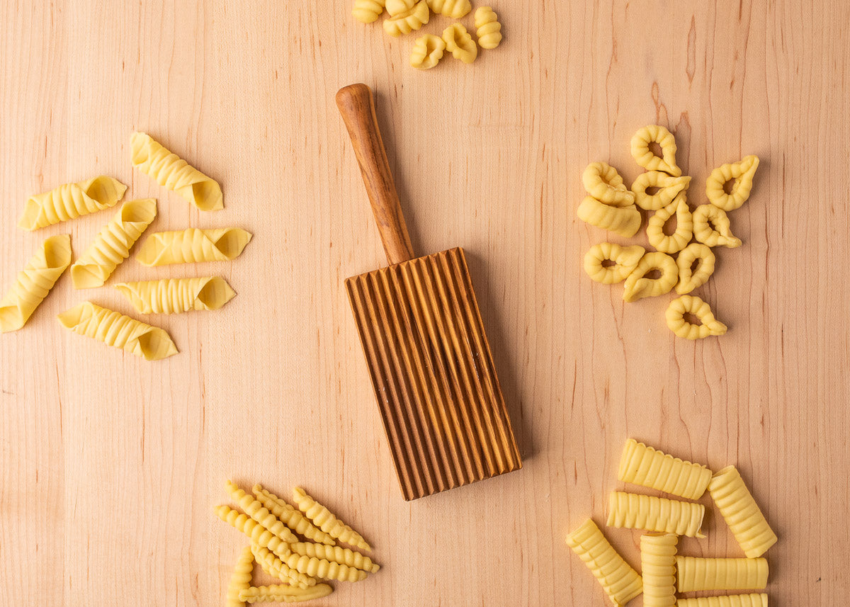 We see a rectangular wooden board with a ridged surface in the center. Around the board are various shapes of pasta, arranged in small piles.