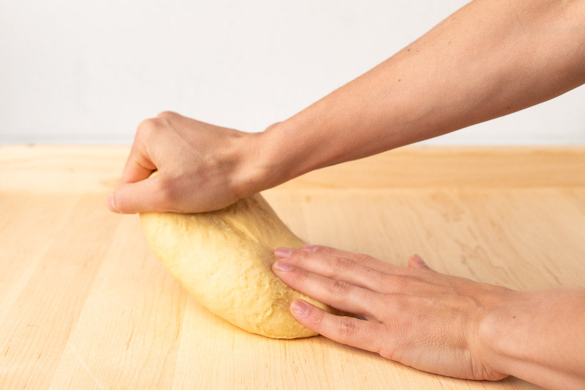 Two hands are stretching a ball of straw-colored pasta dough on a light wooden surface.