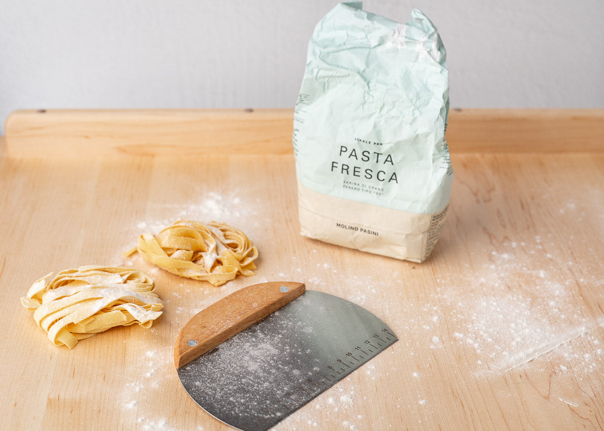 We see two nests of fresh egg taglitelle pasta, a metal bench scraper, and a bag of pasta flour resting on a large, light-colored wooden pasta board.