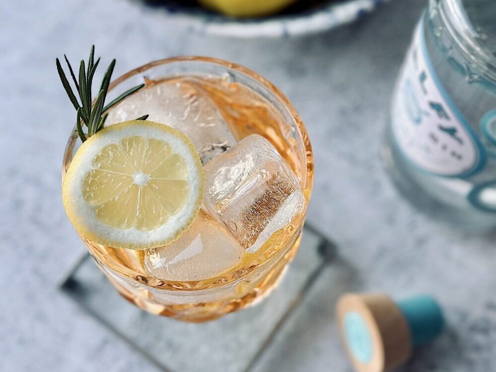 Italian Gin & Tonic - bird's eye view of a blush-colored cocktail in a clear glass. We see a slice of lemon on top and a sprig of rosemary. There is ice in the glass. In the background, we see a bottle of spirits poking through the corner.