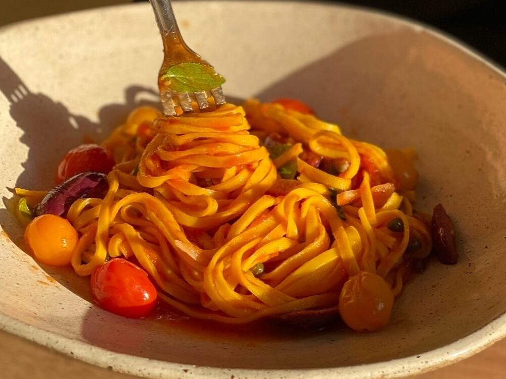 We see a bowl of spaghetti coated in a red tomato sauce. There are a few orange and red cherry tomatoes resting around the spaghetti. A warm sunlight hits the pasta dish, creating dramatic shadows. There is a fork dipping into the spaghetti.