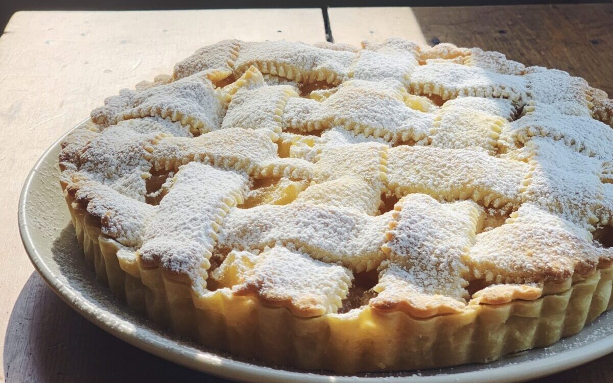 A latticed pie dusted in powdered sugar rests on a wooden surface. The light is warm and sunny.