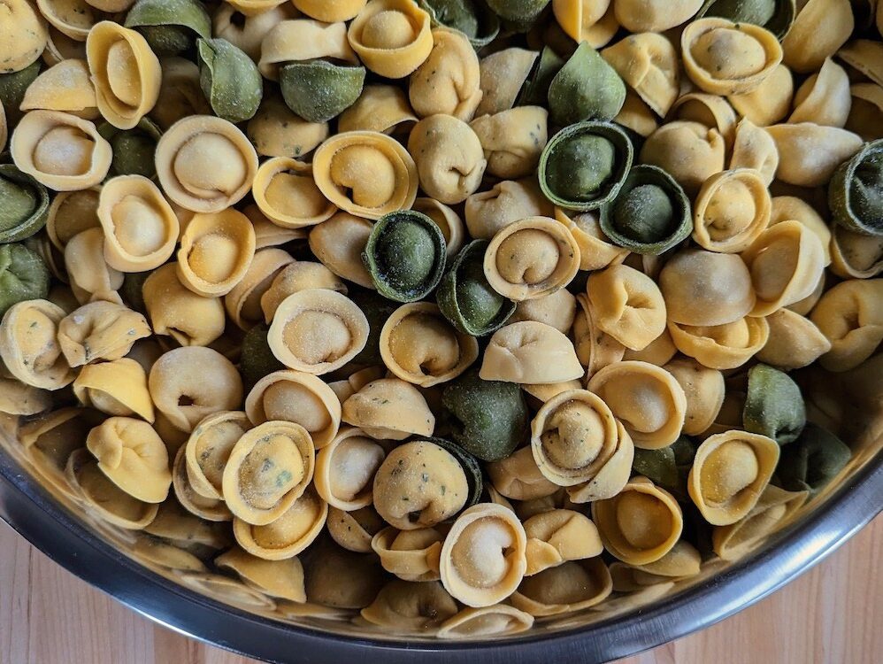 A metal bowl full of green, yellow, and pale yellow stuffed cappelletti pasta. Cappelletti resemble little hats, a round stuffed pasta with a plump center and rimmed edge.