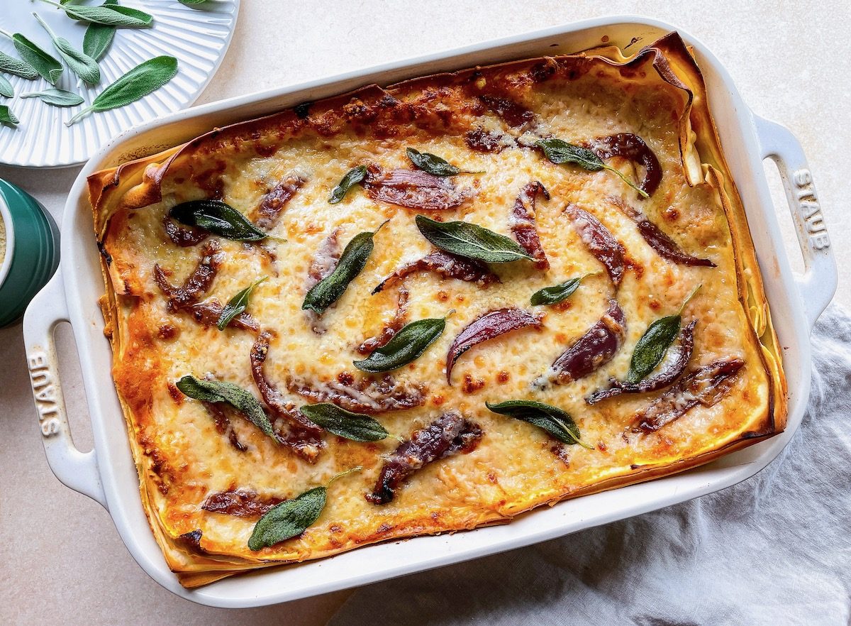Bird's eye view of lasagne in a white ceramic casserole dish. The lasagne is topped with melted cheese, green sage leaves and sliced, cooked red onions. ,