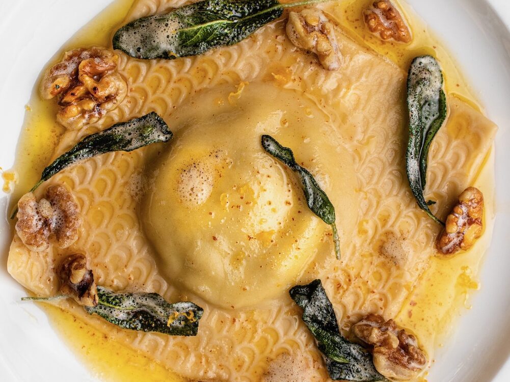 Bird's eye view of a giant square raviolo resting on a white plate. We see an archway pattern on the edges of the ravioli, and a plump center. The raviolo is covered in a brown butter sauce (golden in color) and sprinkled with whole fried sage leaves and 