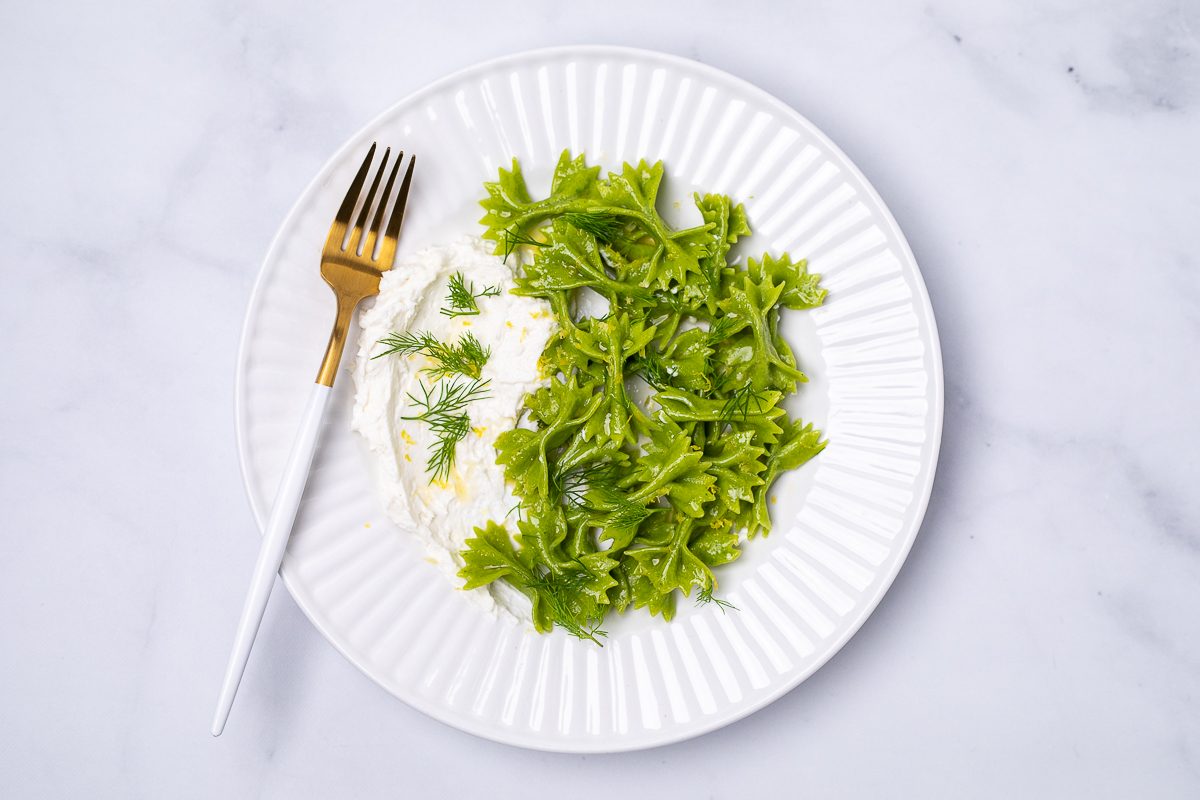 Spinach & dill farfalline pasta on a white plate. On the side we see some whipped goat cheese topped with green dill. There is a gold and white fork resting on the side of the plate.