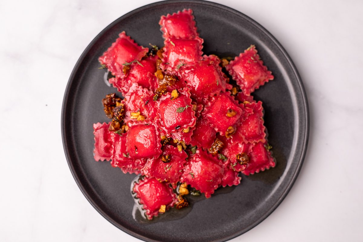 There is a dark grey plate filled with mini square pink ravioli. On top of the ravioli we can see specks of chopped figs, walnuts, and thyme. The plate is sitting on a marble background.