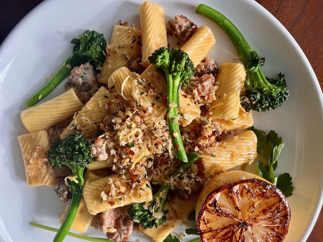 A shallow white bowl holds a mound of homemade rigatoni pasta, cooked broccolini florets, bits of sausage, and toasted breadcrumbs. In the bottom right corner of the bowl, sits a lemon half, with caramelized edges.