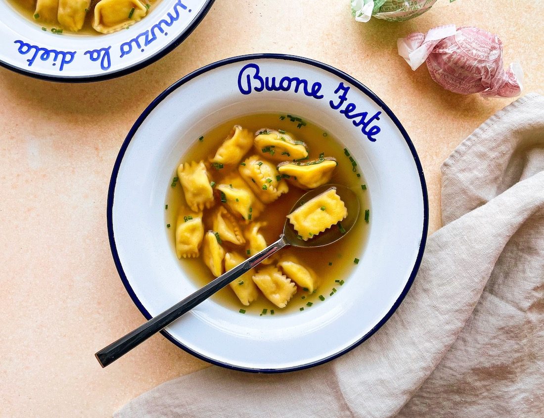 Agnolotti di plin pasta in a broth served in a white bowl that reads Buone Feste in blue