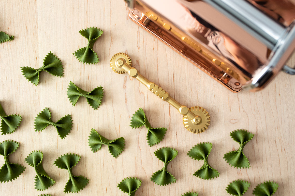 Green bowtie pasta is scattered across a light wooden surface. We see a brass pasta wheel, with a wheel on each end. In the top right corner, there is a copper pasta machine peeking through.