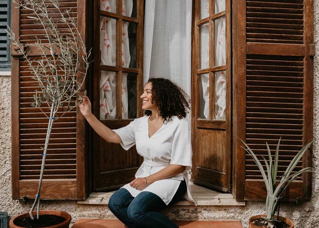Skyler Mapes of EXAU Olive Oil sitting in front of wooden french doors. Her hand is reaching out to touch a potted olive tree.