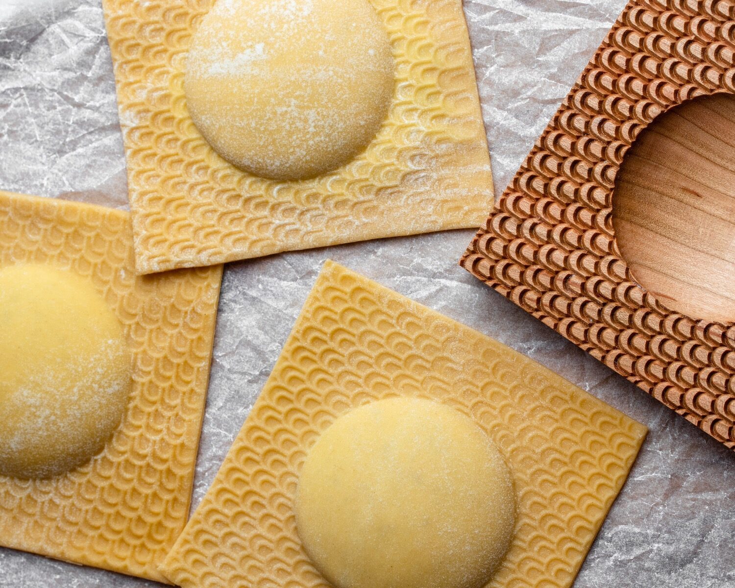 Three big ravioli sit on crumpled parchment paper. We see the edge of a wooden ravioli mold off to the right.