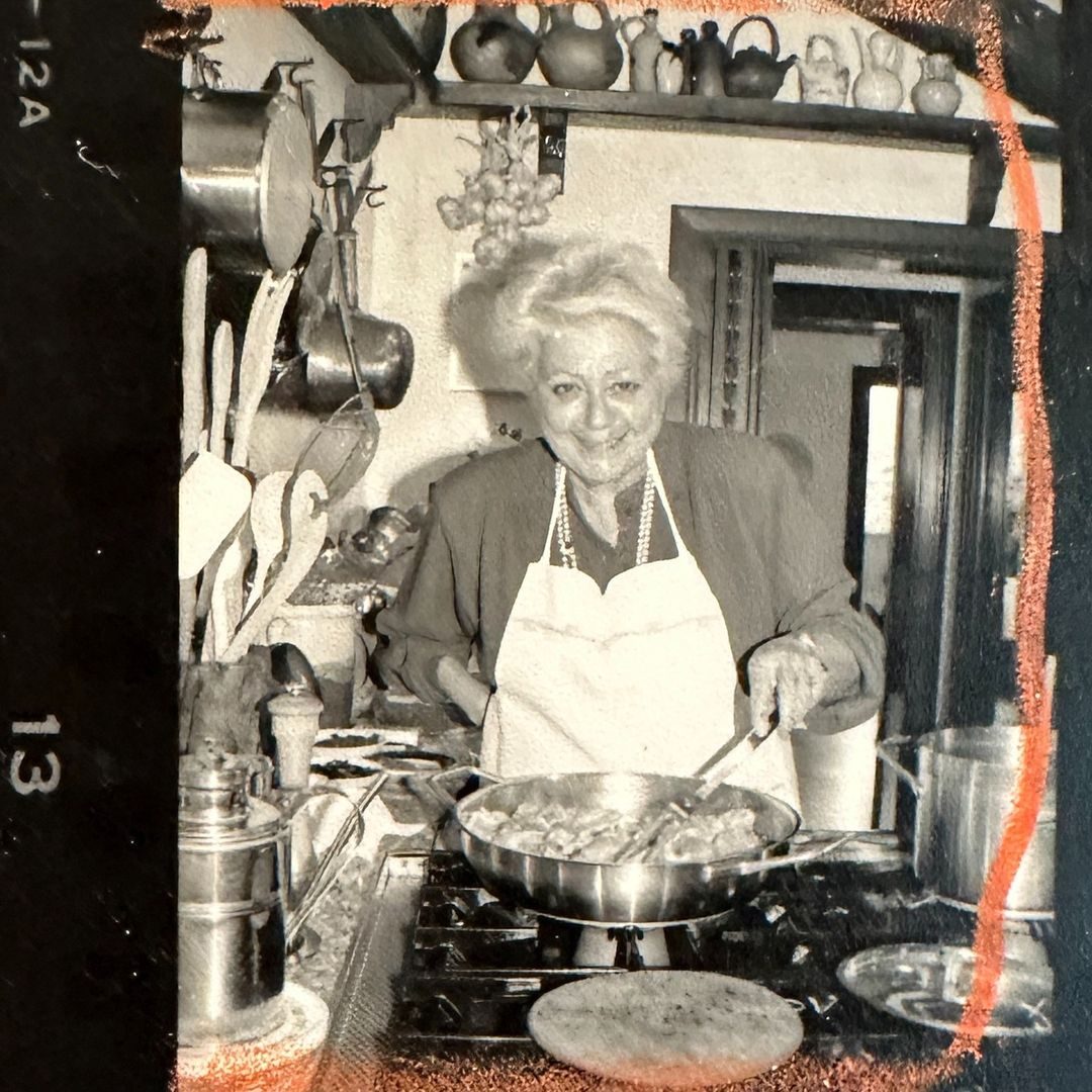 Old black and white photo of Marcella Hazan stirring a pan over a stove. She's looking up at the camera and smiling. Photo taken by a student, of a class taught by Marcella Hazan in Venice in the 1980s.