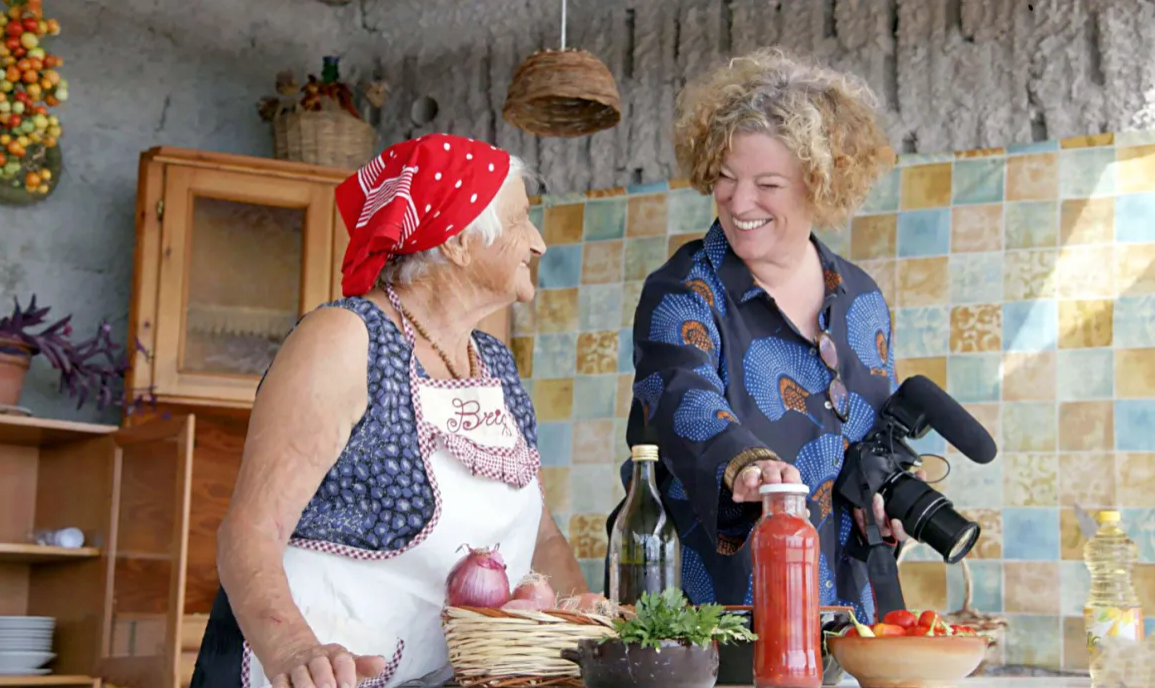 An elderly woman in a red bandana and apron looks to her left towards Vicky Bennison, a tall woman with curly hair. Vicky is holding a video camera with a microphone. The two women are smiling at one another.