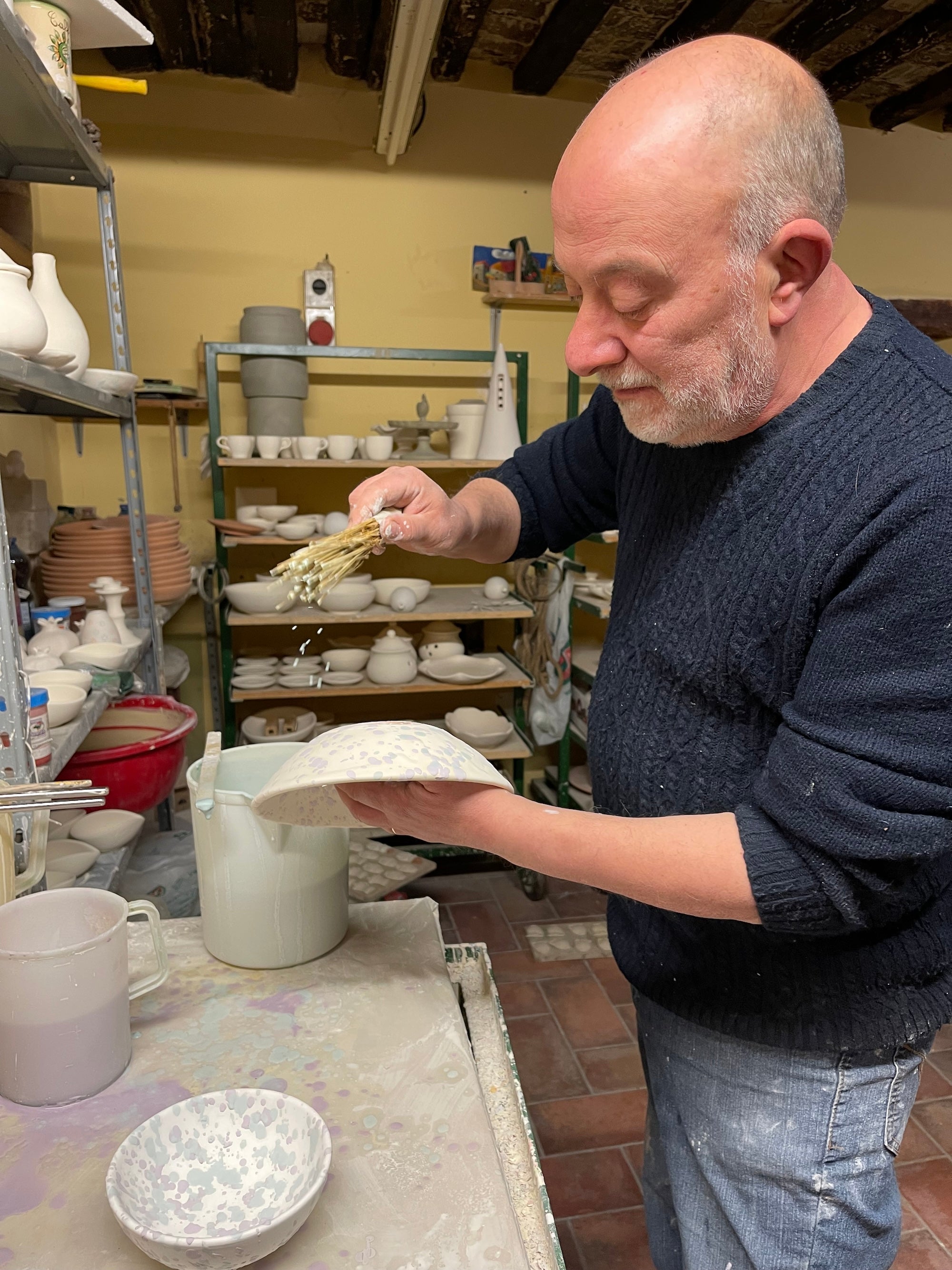 A man with a gray beard is holding a broom-like brush. The brush is dripping paint over a bowl. We can see in the background that there is some kind of pottery studio.