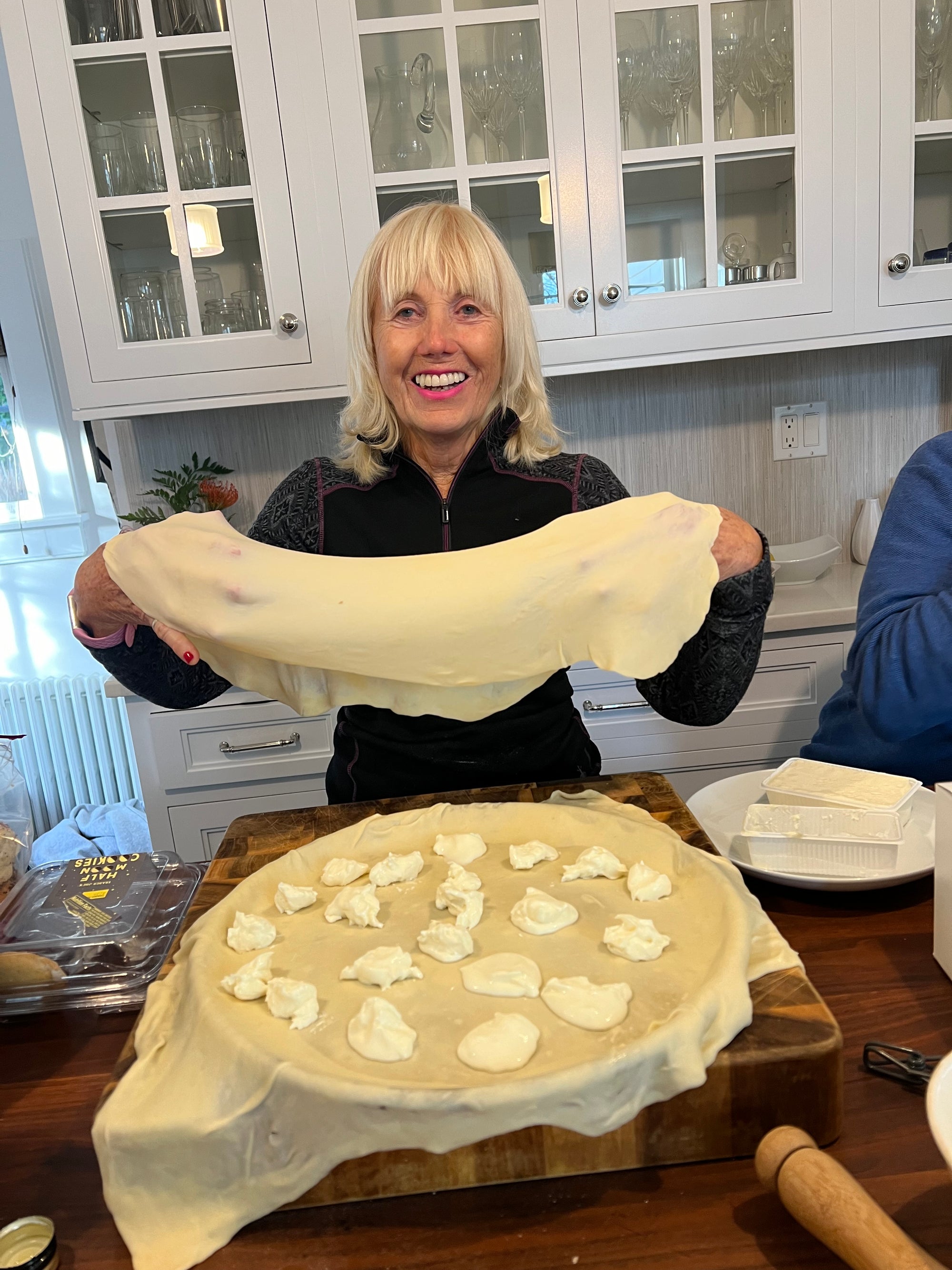 A woman is holding up a thin piece of dough, stretched out between her hands. Below it is a pan covered in another thin sheet of pastry, and dotted with a white soft cheese. The woman is smiling.