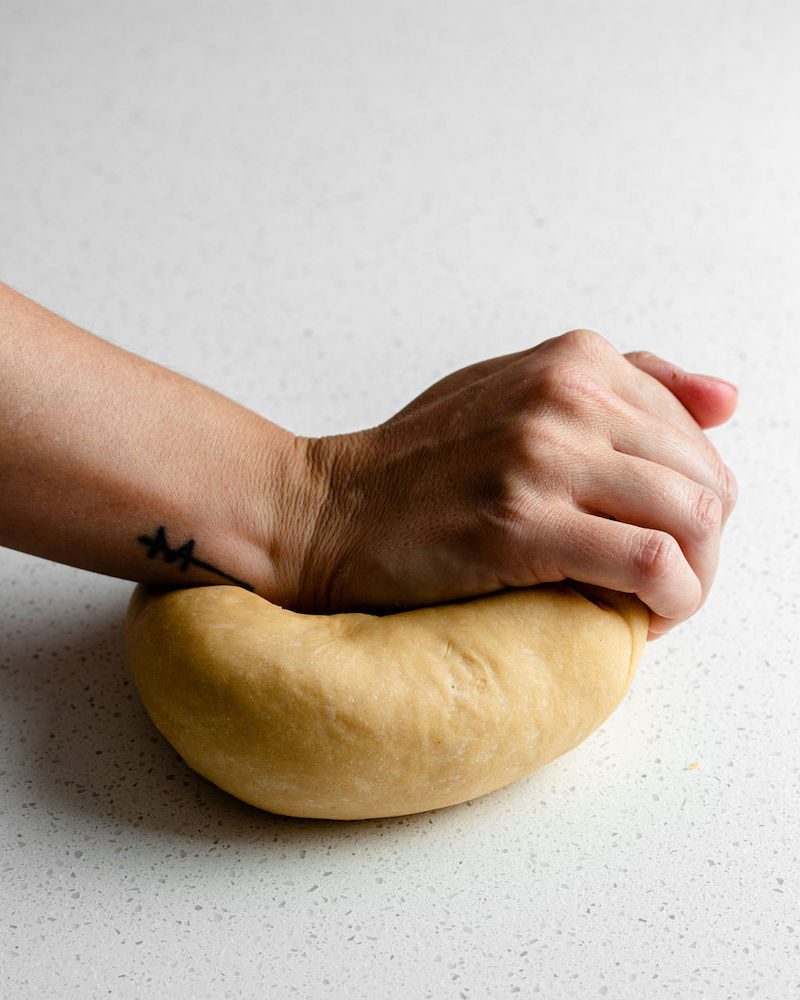 Side angle view of a hand is kneading a pale yellow pasta dough on a white speckled surface.